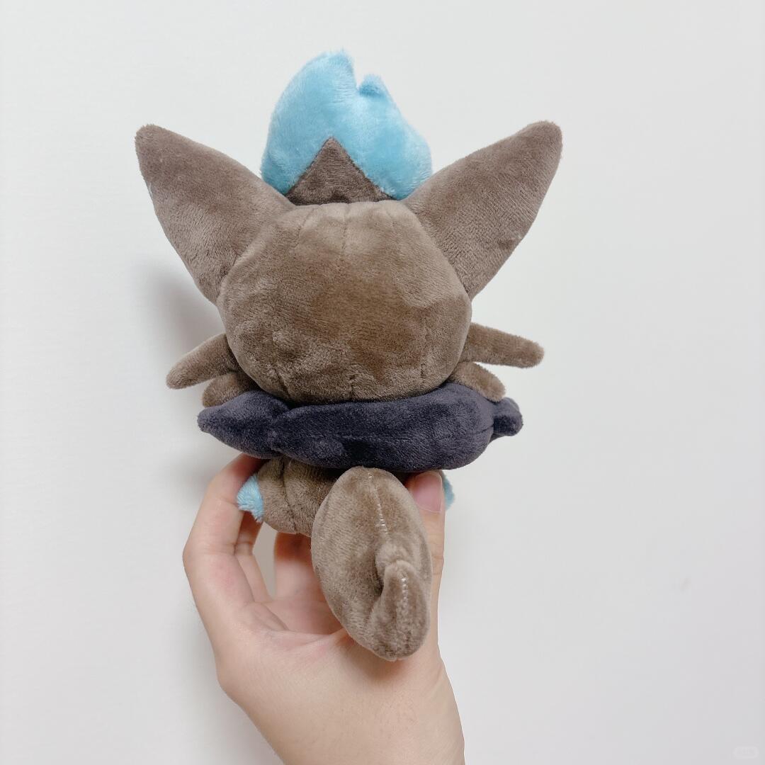 A zorua plush toy held in a person's hand against a plain light background. The toy appears to be a small brown animal character with large pointed ears, a light blue tuft on top, and a dark navy ring around the middle.