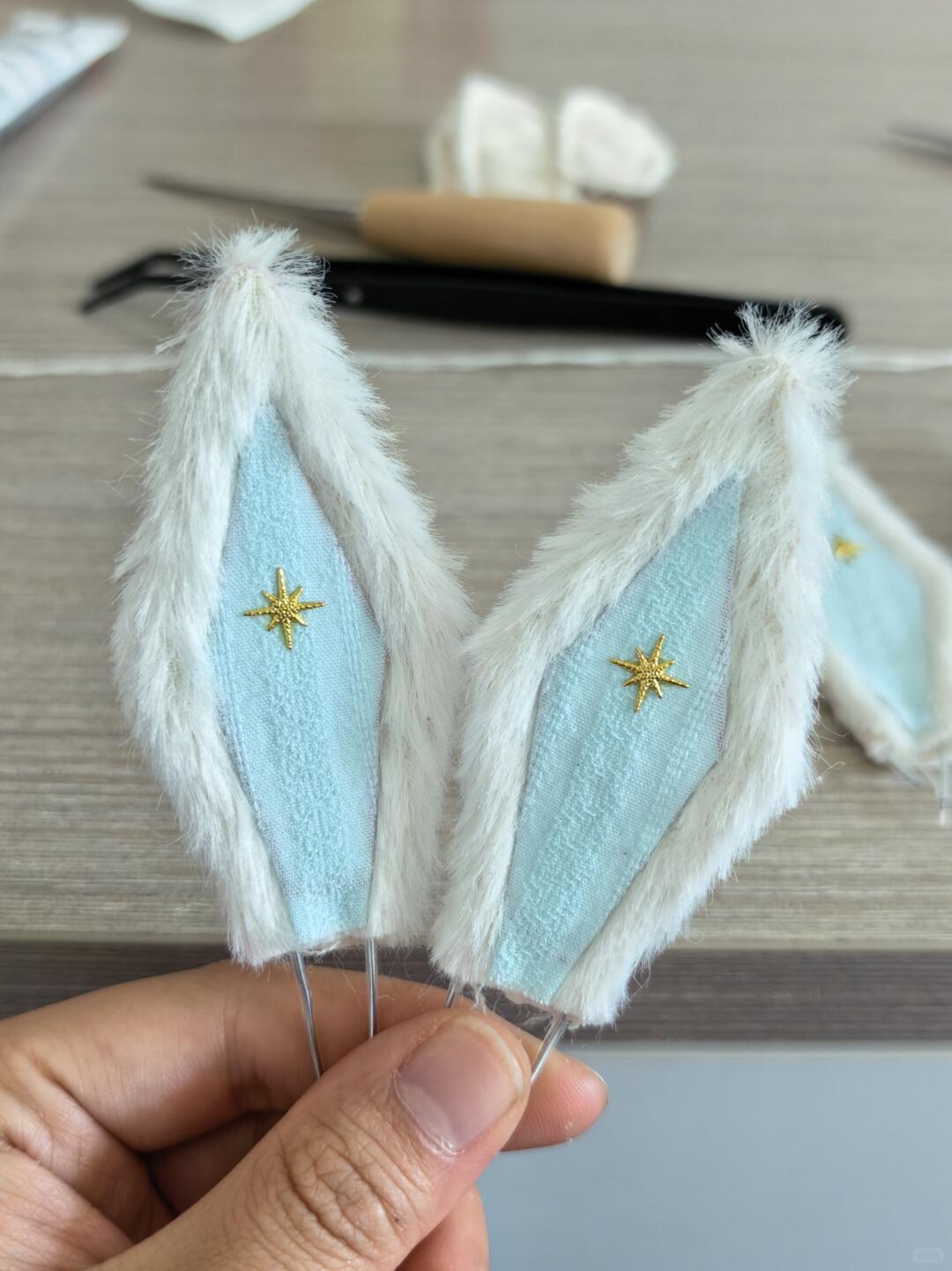 A person's hand holding a pair of handmade costume or doll ears. The ears feature fluffy white faux fur trim, a light blue fabric center, and a small gold embroidered star on each side, set against a blurred wooden table background.