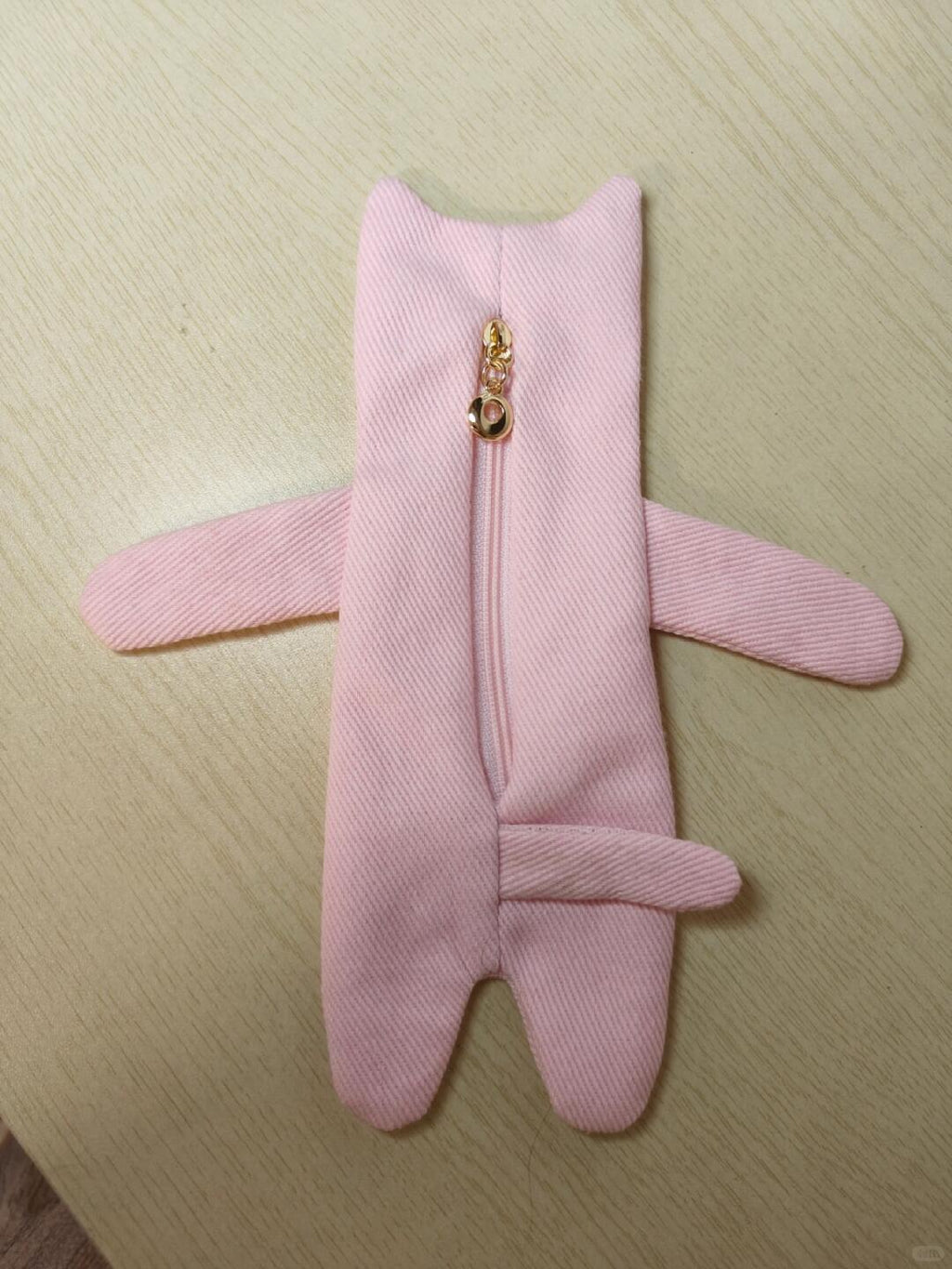 A pink fabric pouch shaped like a small character or animal, with long arms, a short tail, and a gold zipper running down the center, laid flat on a light wood surface.