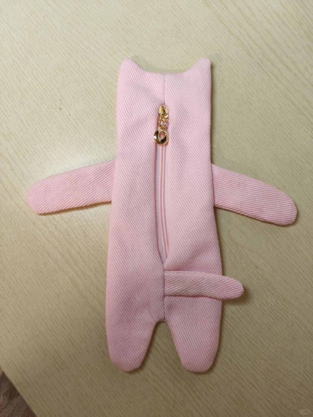 A pink fabric pouch shaped like a small character or animal, with long arms, a short tail, and a gold zipper running down the center, laid flat on a light wood surface.