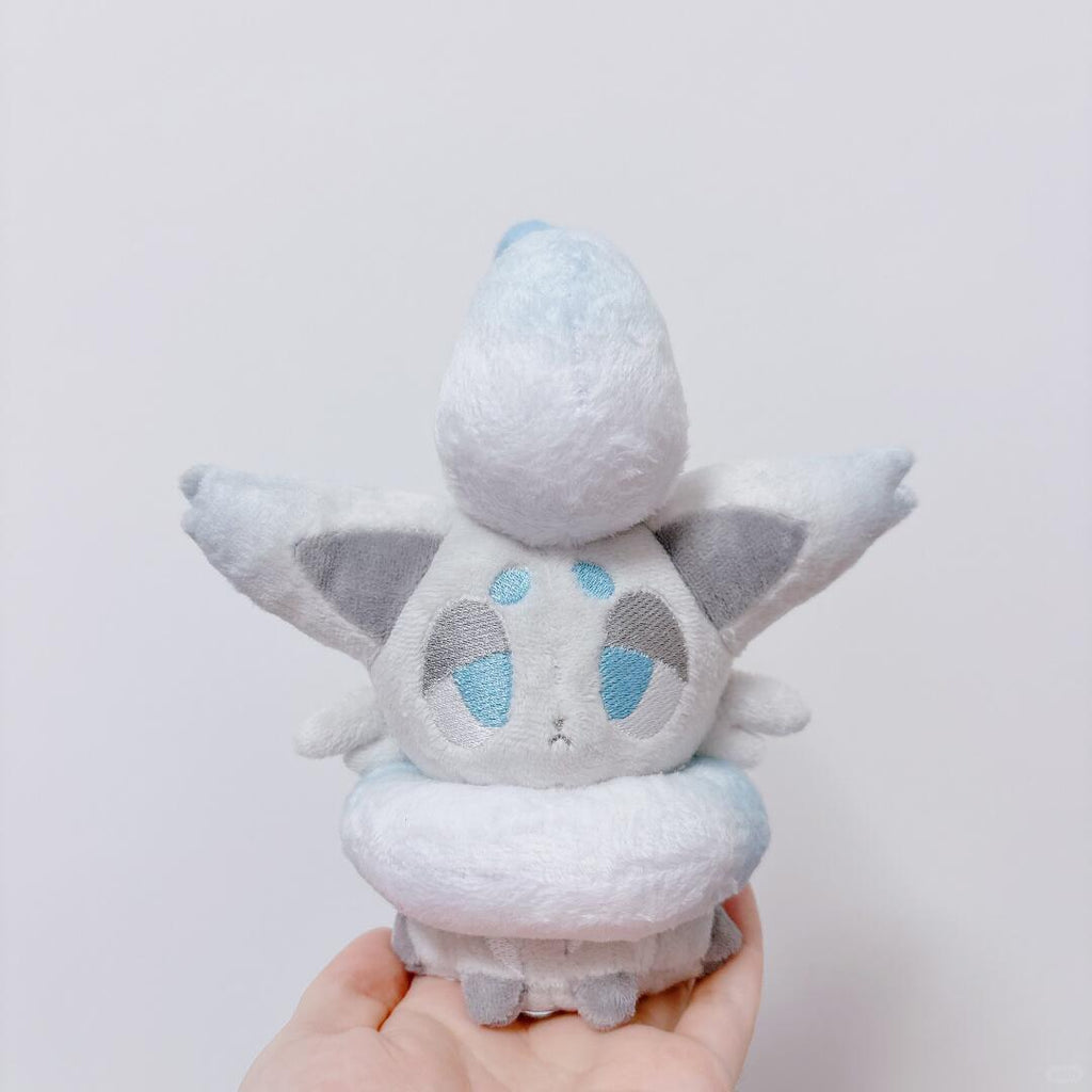 A small zorua plush toy of a cute, fluffy white creature with blue and gray embroidered eyes and wings, held in a person's hand against a plain light gray background.