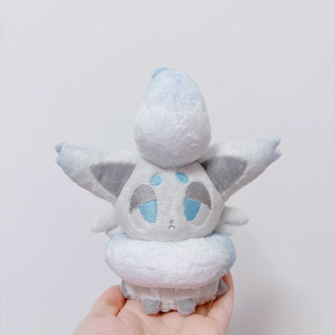 A small zorua plush toy of a cute, fluffy white creature with blue and gray embroidered eyes and wings, held in a person's hand against a plain light gray background.