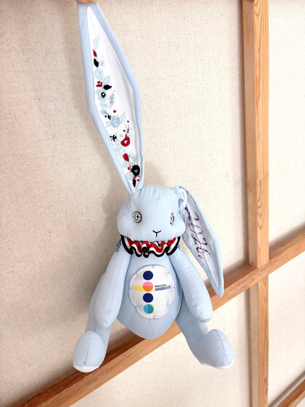 Light blue denim bunny plush hanging from an embroidered ear in front of a wooden display frame.