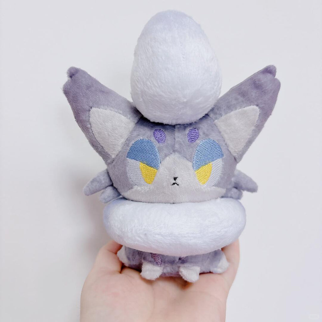 A small zorua plush toy with a gray and white body, blue and yellow embroidered eyes, and a white rounded top, being held in a person's hand against a plain light background.