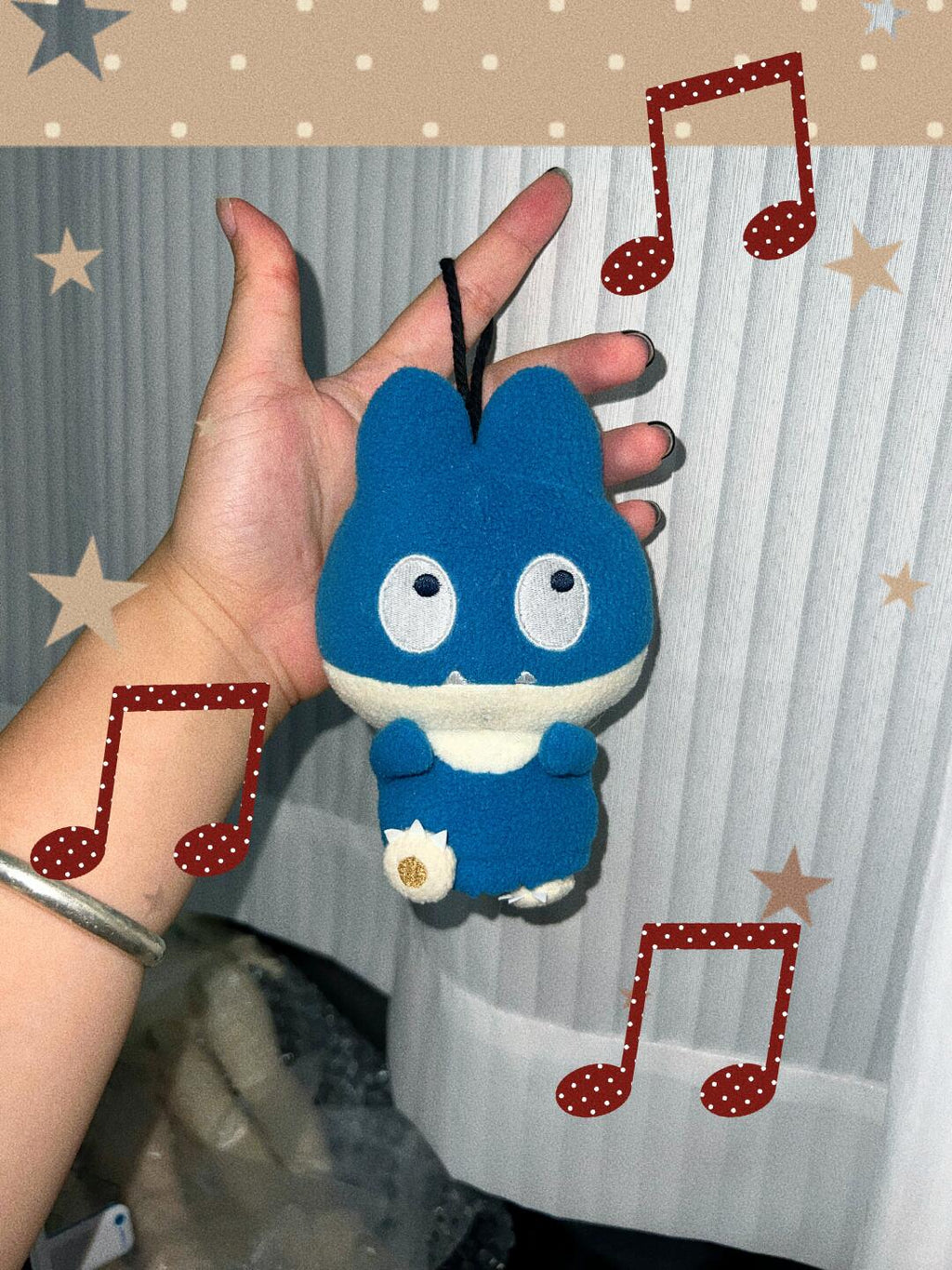 A hand holds a small blue and white plush snorlax character keychain or bag charm in front of a white ribbed curtain, with decorative music note and star graphics overlaid around the photo.