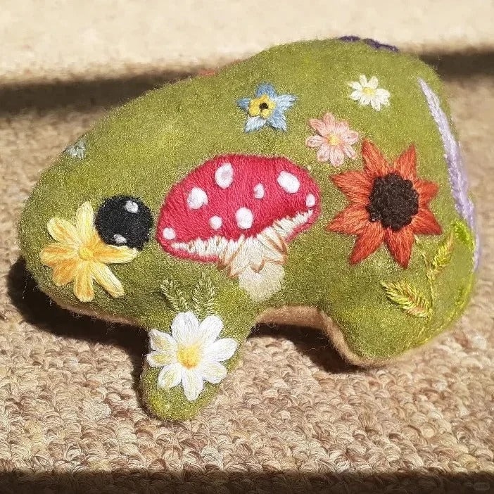 Green fabric frog decorated with embroidered flowers and a red mushroom with white spots.
