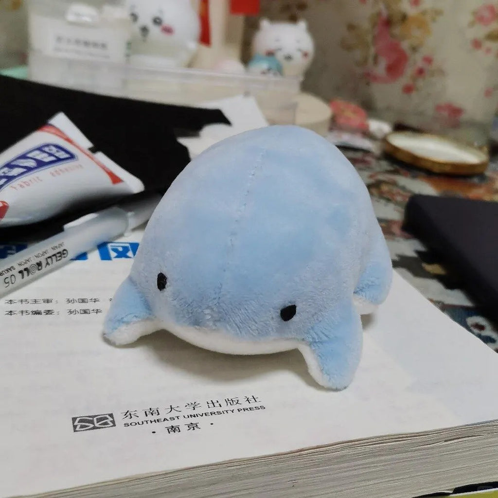 Pale blue frog plush with embroidered black eyes and a white belly resting on a book.
