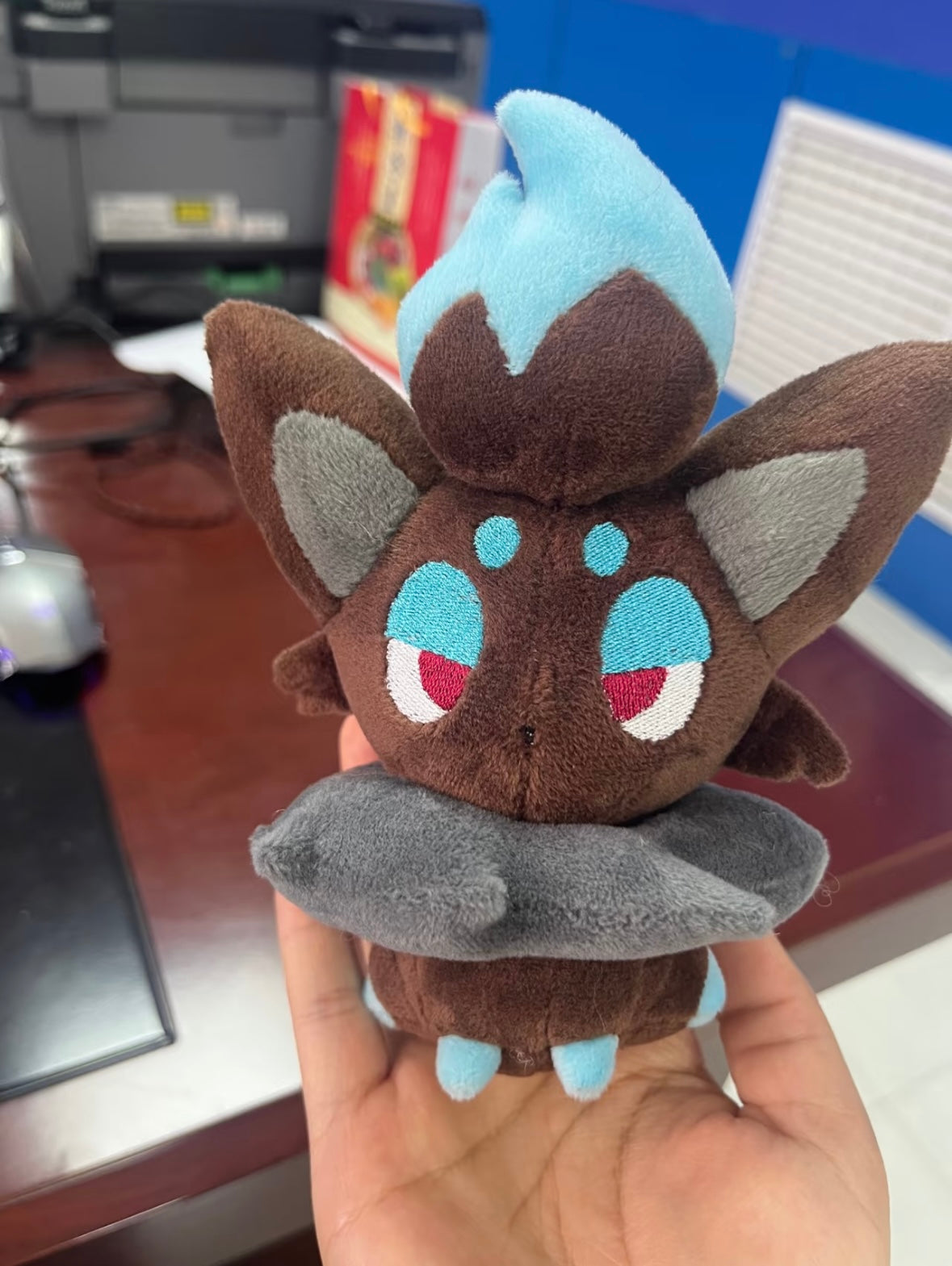 A small plush toy of a brown cartoon creature Zorua with large pointed ears, light blue and gray accents, and embroidered eyes, being held in a person's hand in an office setting.
