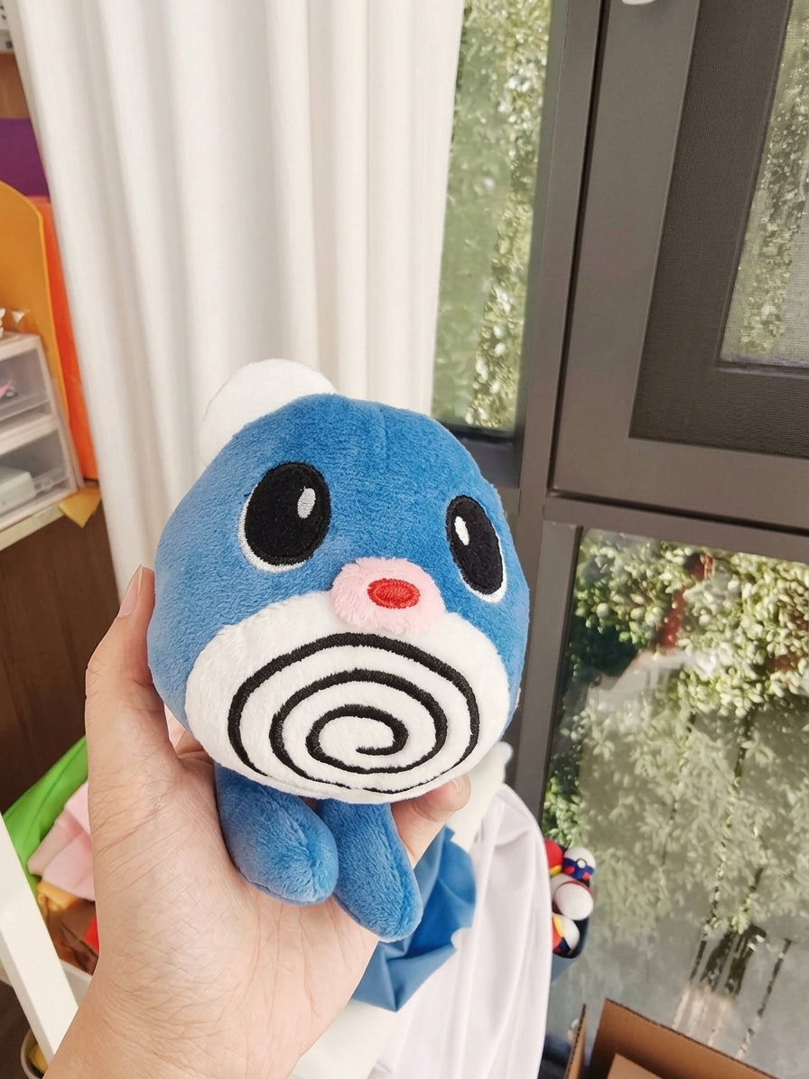 A blue and white plush Poliwag toy with a spiral face design is held in a person's hand in front of a window with white curtains.