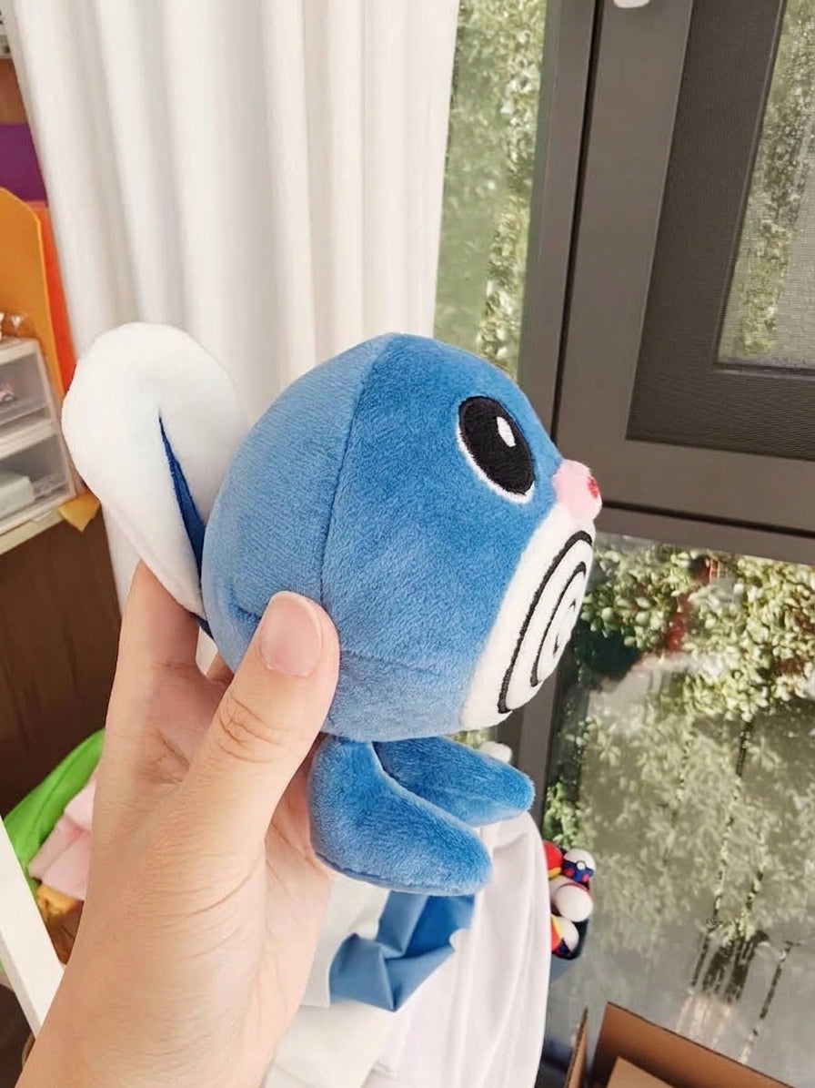A person's hand holding a blue and white Poliwag  plush toy with a large black eye and pink nose, photographed indoors near a window.