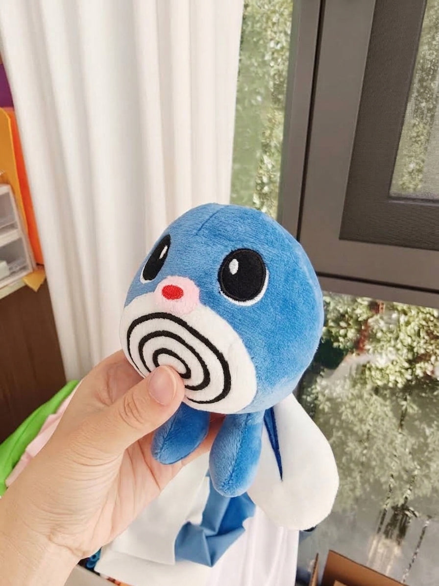 A blue and white Poliwag plush toy with a spiral face design is held up by a hand in front of a window with white curtains.