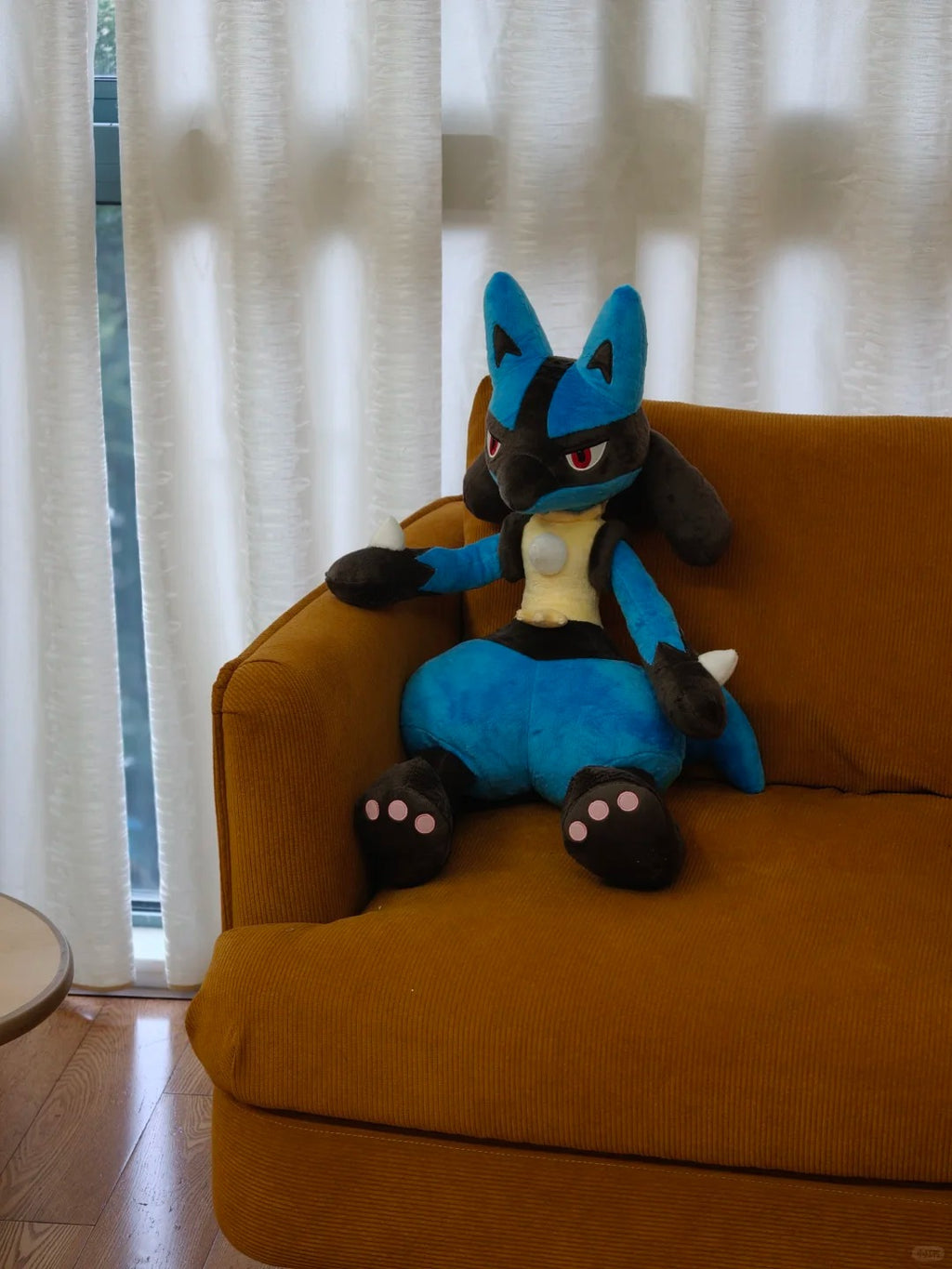 A blue and black pokemon lucario plush toy with red eyes sits on an orange-brown armchair in front of white curtains.