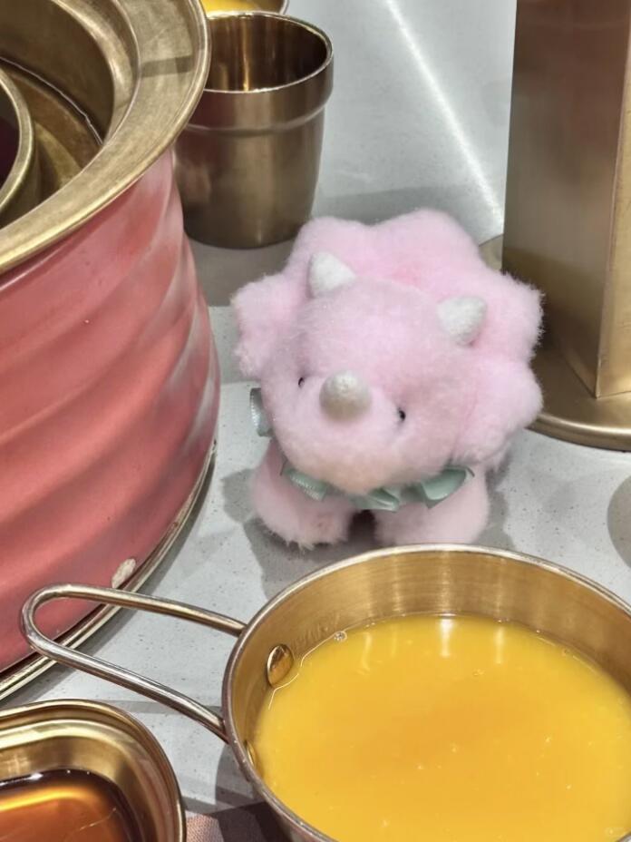 Pink plush dinosaur toy with pearl-like buttons next to a pot of yellow liquid on a metallic surface.