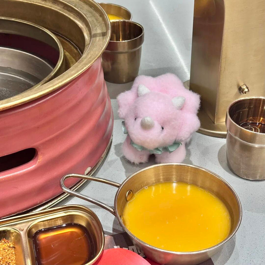 Pink ceramic pot with gold trim, small pink plush dinosaur toy, and metal containers on a light gray surface.