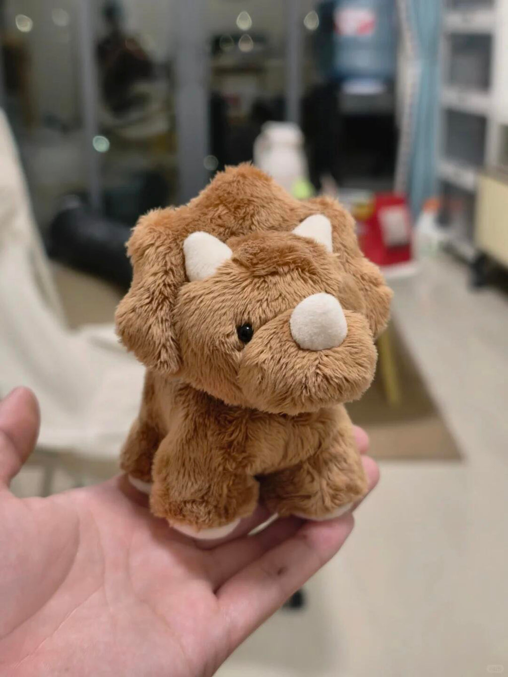 Small brown dinosaur plush toy held in a hand with a blurred indoor background