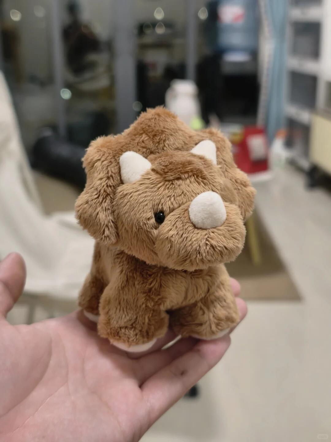 Small brown dinosaur plush toy held in a hand with a blurred indoor background