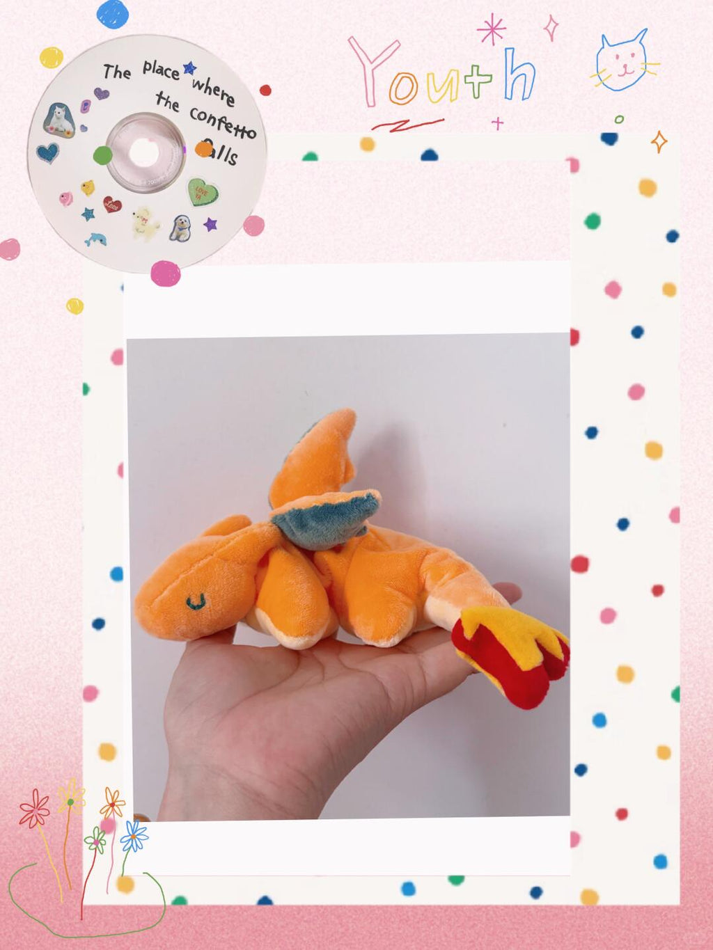 A small orange charizard plush toy with red and yellow flame details and a blue wing is being held on a person's hand, centered within a white-bordered photo frame on a pink polka-dot background. Decorative text and doodles appear around the frame.