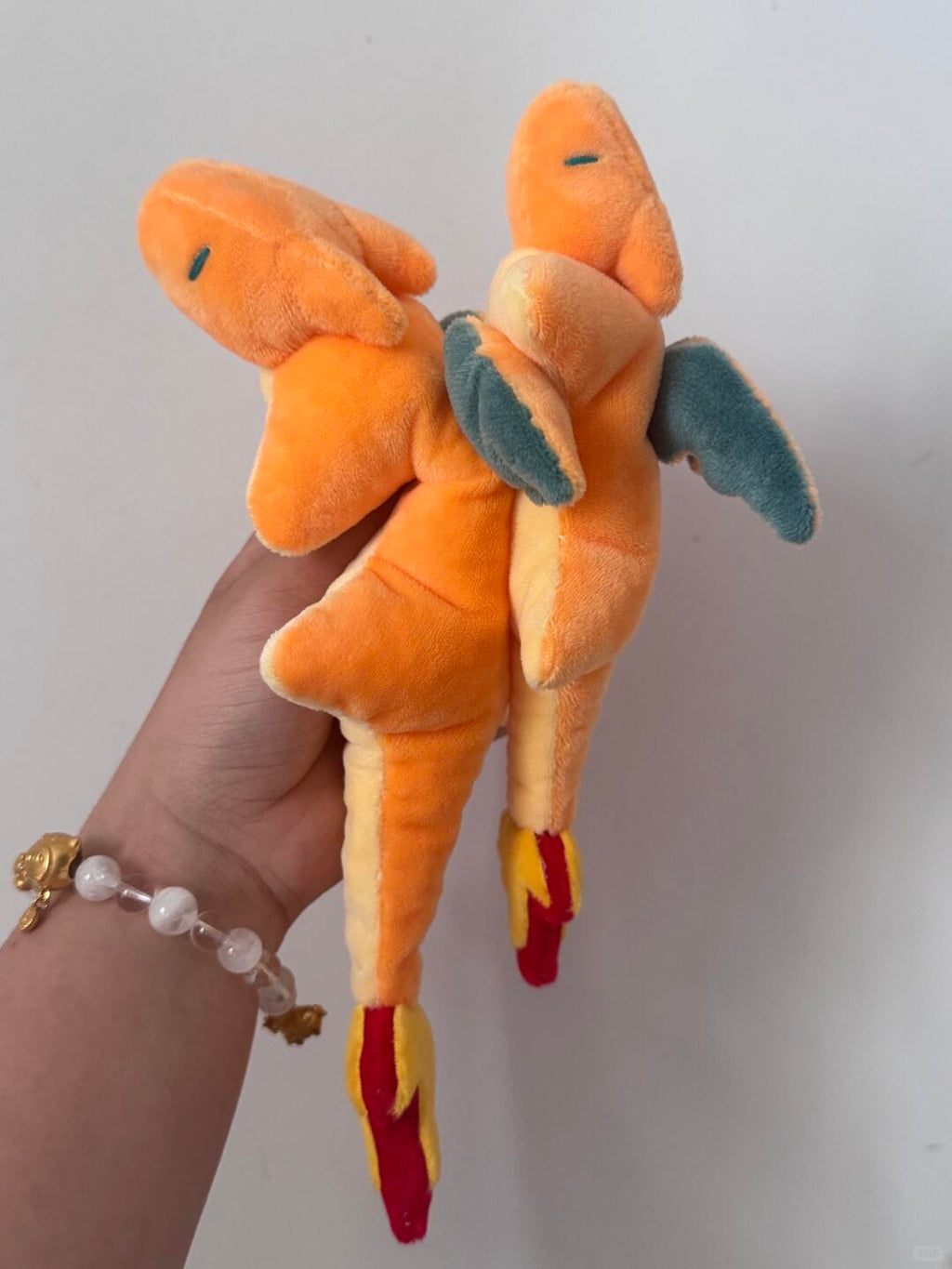A plush toy resembling Charizard, held in a person's hand against a plain light gray wall. The toy is orange with yellow underbelly, blue wings, and red-yellow flame details.