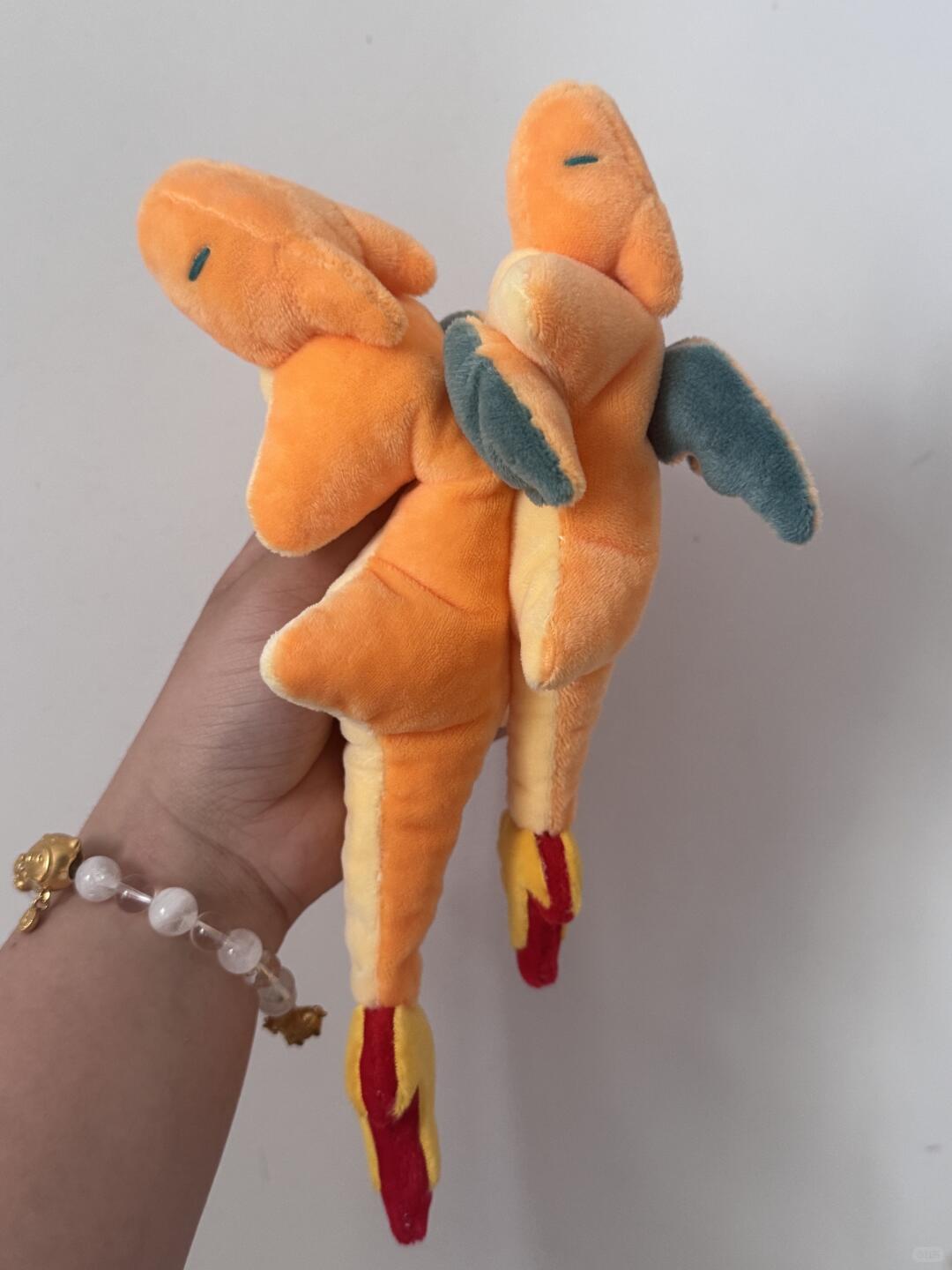 A plush toy resembling Charizard, held in a person's hand against a plain light gray wall. The toy is orange with yellow underbelly, blue wings, and red-yellow flame details.