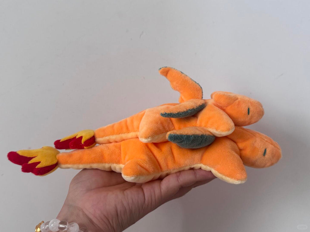An orange plush toy resembling a dragon or fire-breathing charizard creature, held in a person's hand against a plain light gray wall.