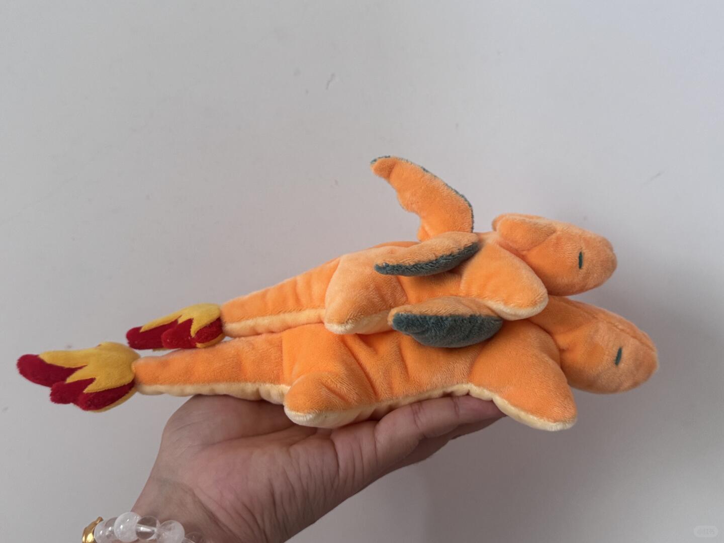 An orange plush toy resembling a dragon or fire-breathing charizard creature, held in a person's hand against a plain light gray wall.