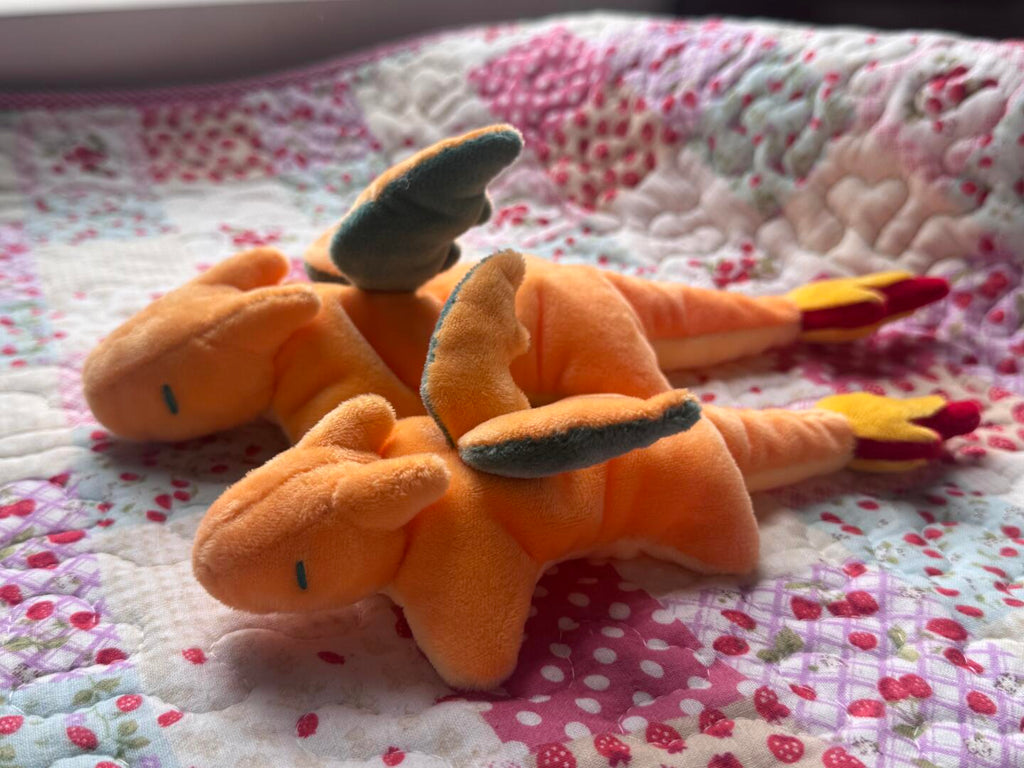 Two orange plush toy charizard characters with gray wings and red-yellow tails resting on a pink and white patterned quilted blanket.