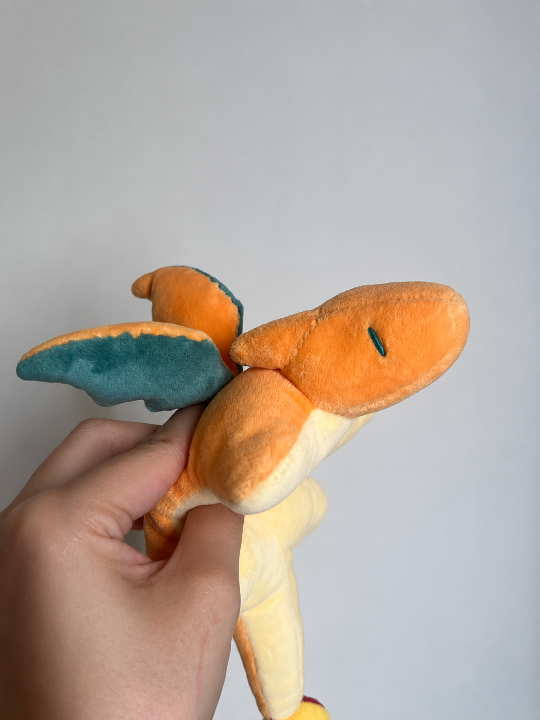 A hand holding a plush toy of Charizard, showing its orange body, yellow underside, and teal wings against a plain light gray background.