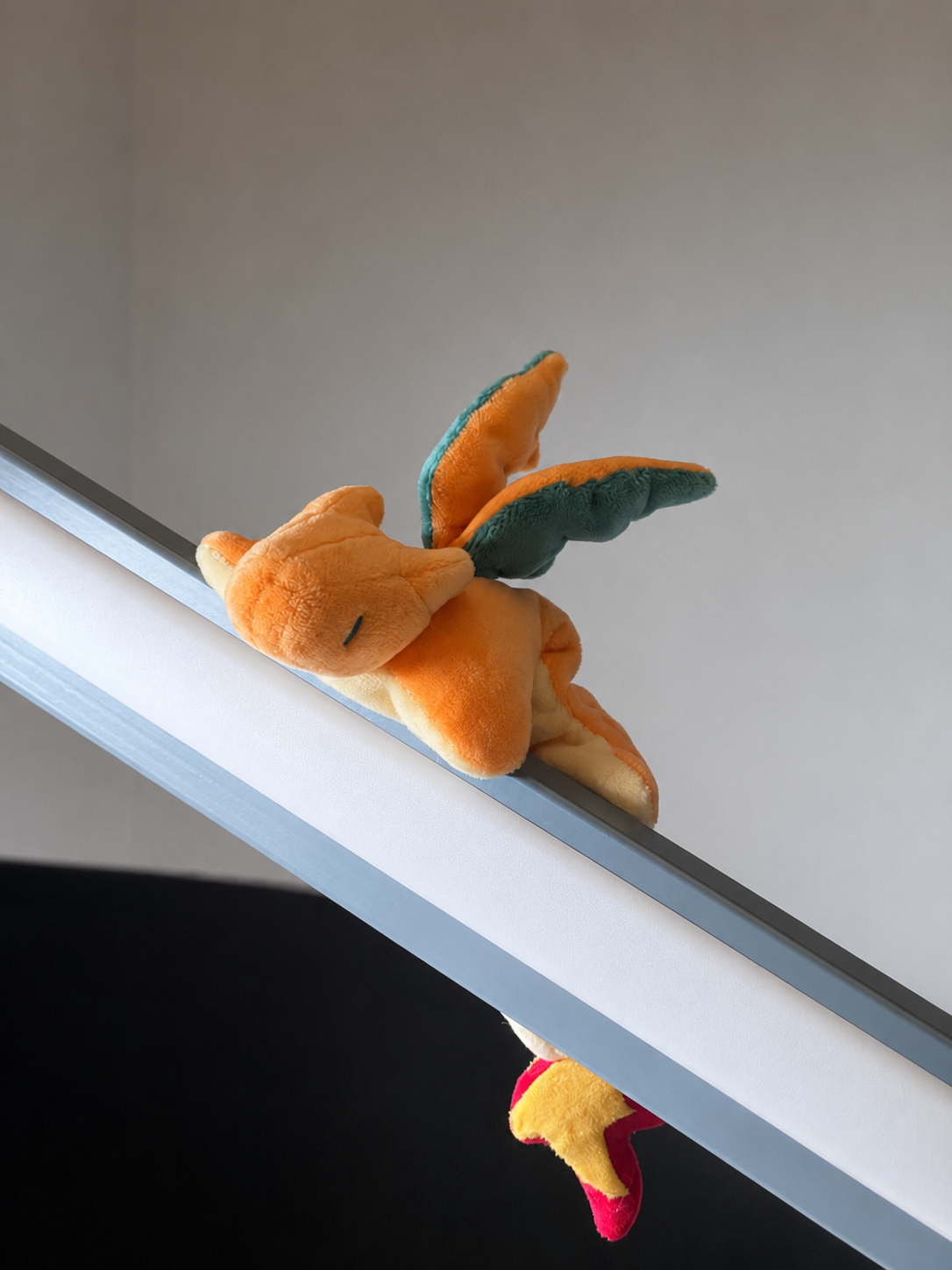 A small orange charizard plush toy with teal wings and a yellow-and-pink lower tail, photographed resting on a white ledge against a plain light wall.