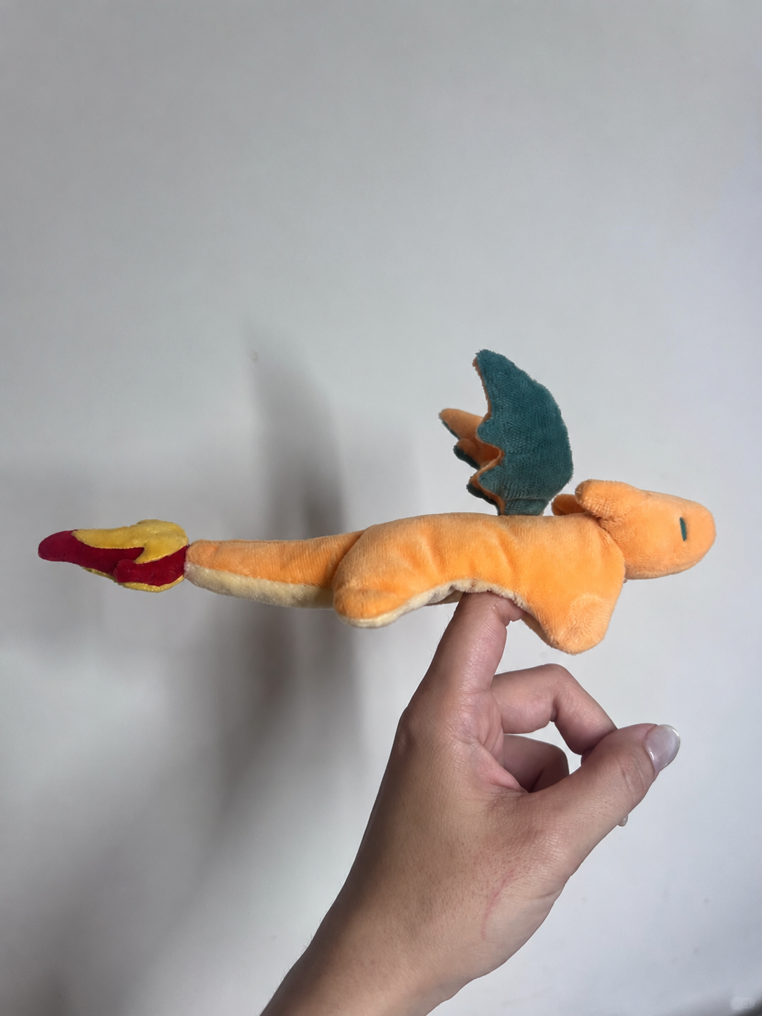 A plush toy of Charizard, an orange dragon with a teal wing and red-yellow flame tail, held by a person's hand against a plain light gray background.