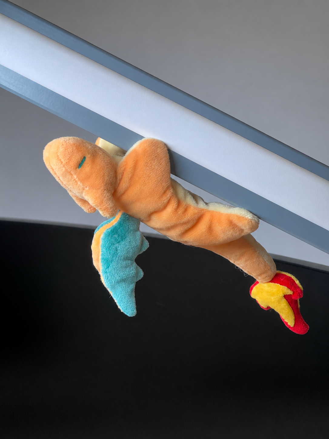 A charizard plush toy shaped like a small orange creature with a blue wing and red-and-yellow feet, hanging against a gray and black background.
