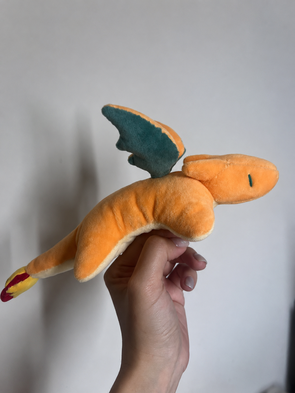 An orange plush toy charizard dragon with teal wings and a red-tipped tail, held in a person's hand against a plain light gray wall.