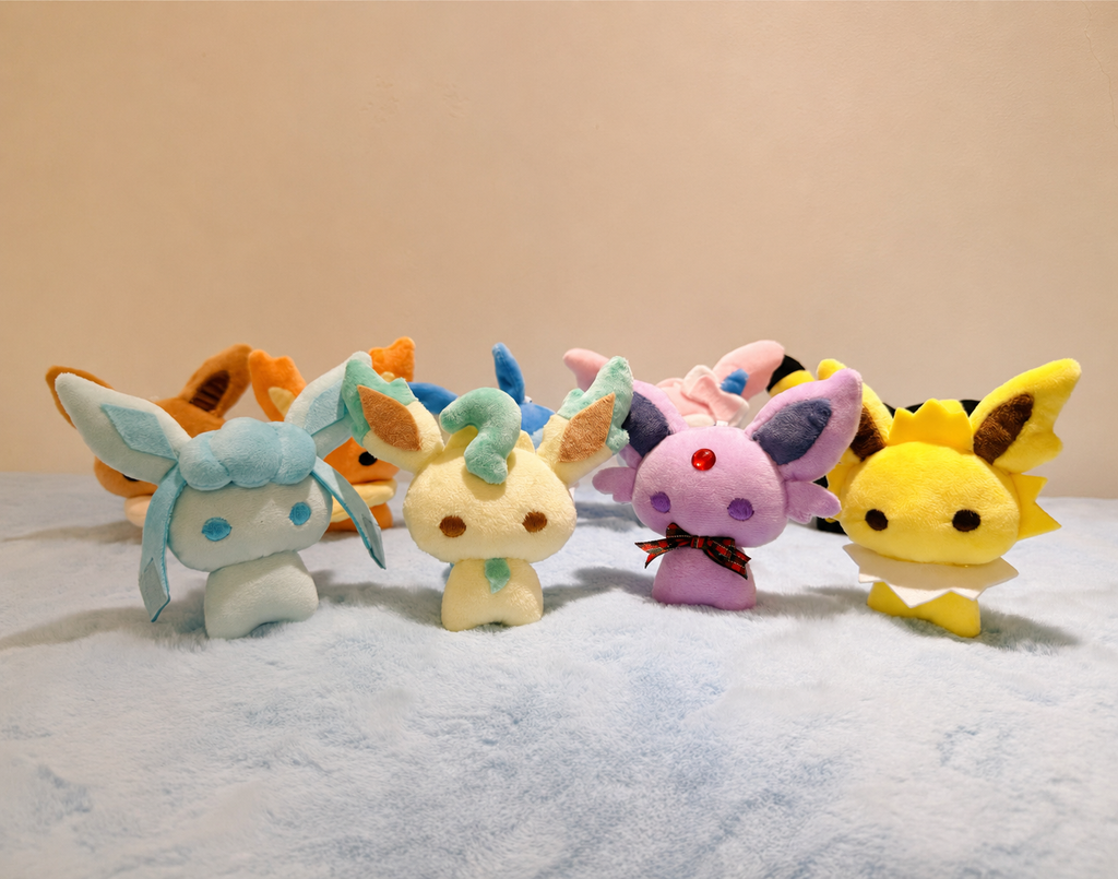 Handmade Pokémon-inspired Eeveelution plush toys including Glaceon, Leafeon, Espeon, Jolteon, and Eevee, displayed together on a soft blanket. Cute handmade plush sewing pattern sample.