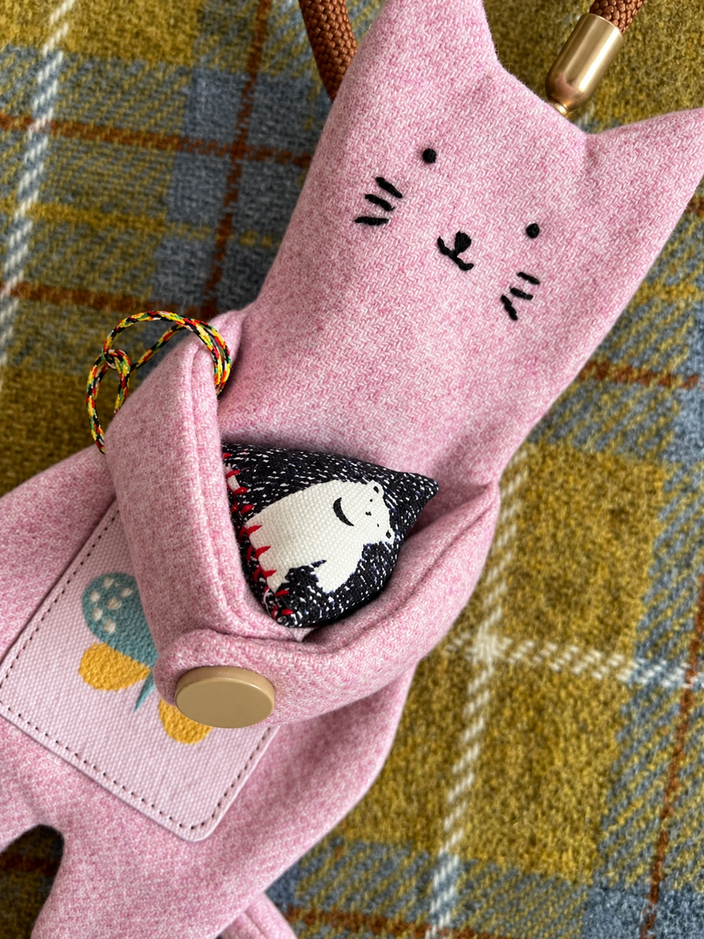 A pink felt cat-shaped pouch or organizer with embroidered cat face details, a button closure, and a small black-and-white cat-shaped fabric insert peeking out, photographed on a yellow and blue plaid fabric background.