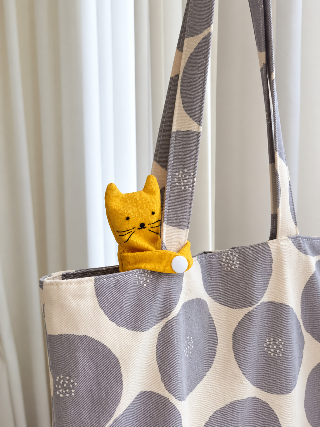 A close-up of a tote bag with a cream and gray polka dot pattern, featuring a small yellow cat-shaped fabric accessory attached near the top zipper.
