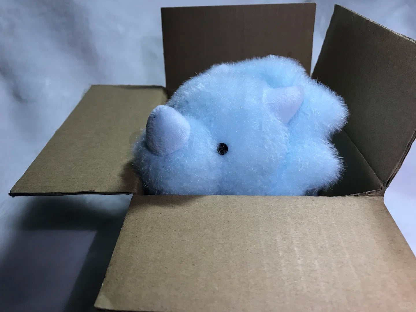 Blue dinosaur plush toy inside a cardboard box on a light gray background