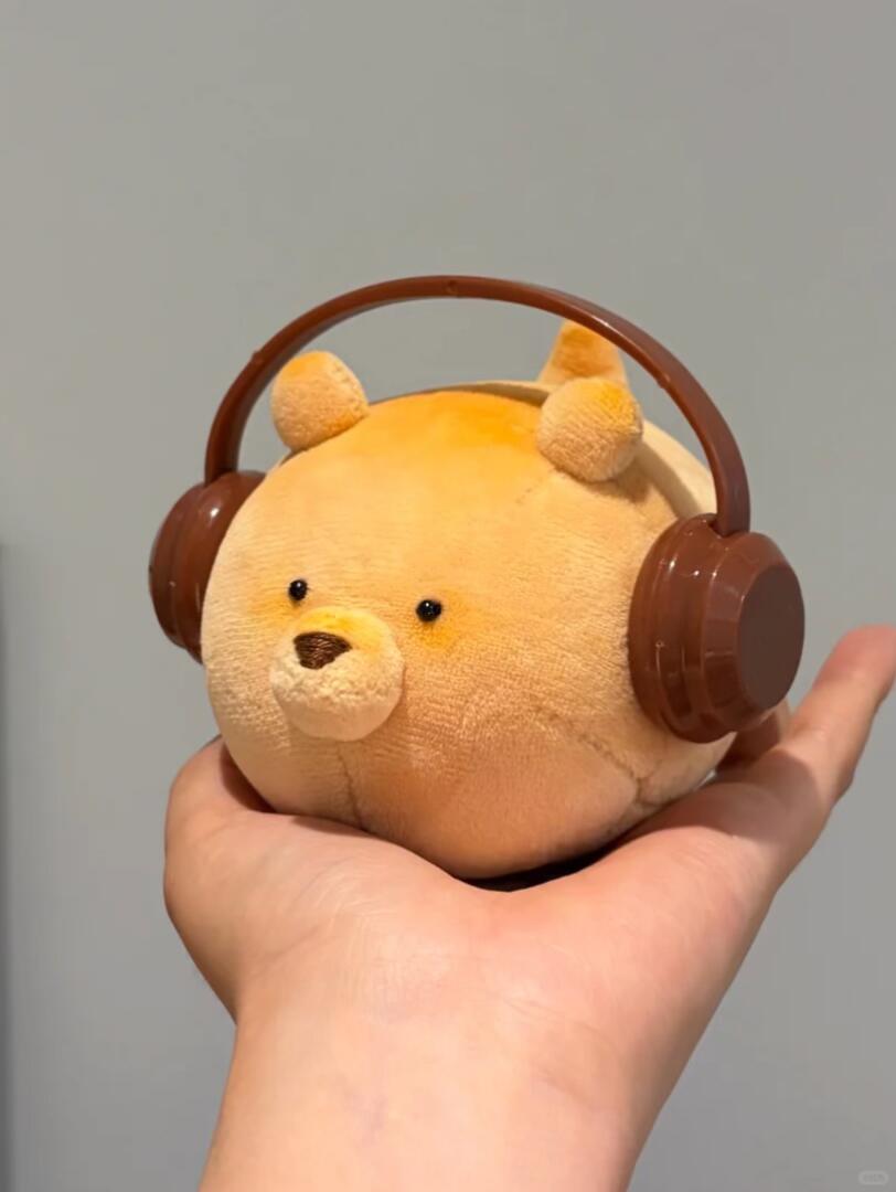 Plush toy resembling a dog with brown headphones held in a hand against a gray background