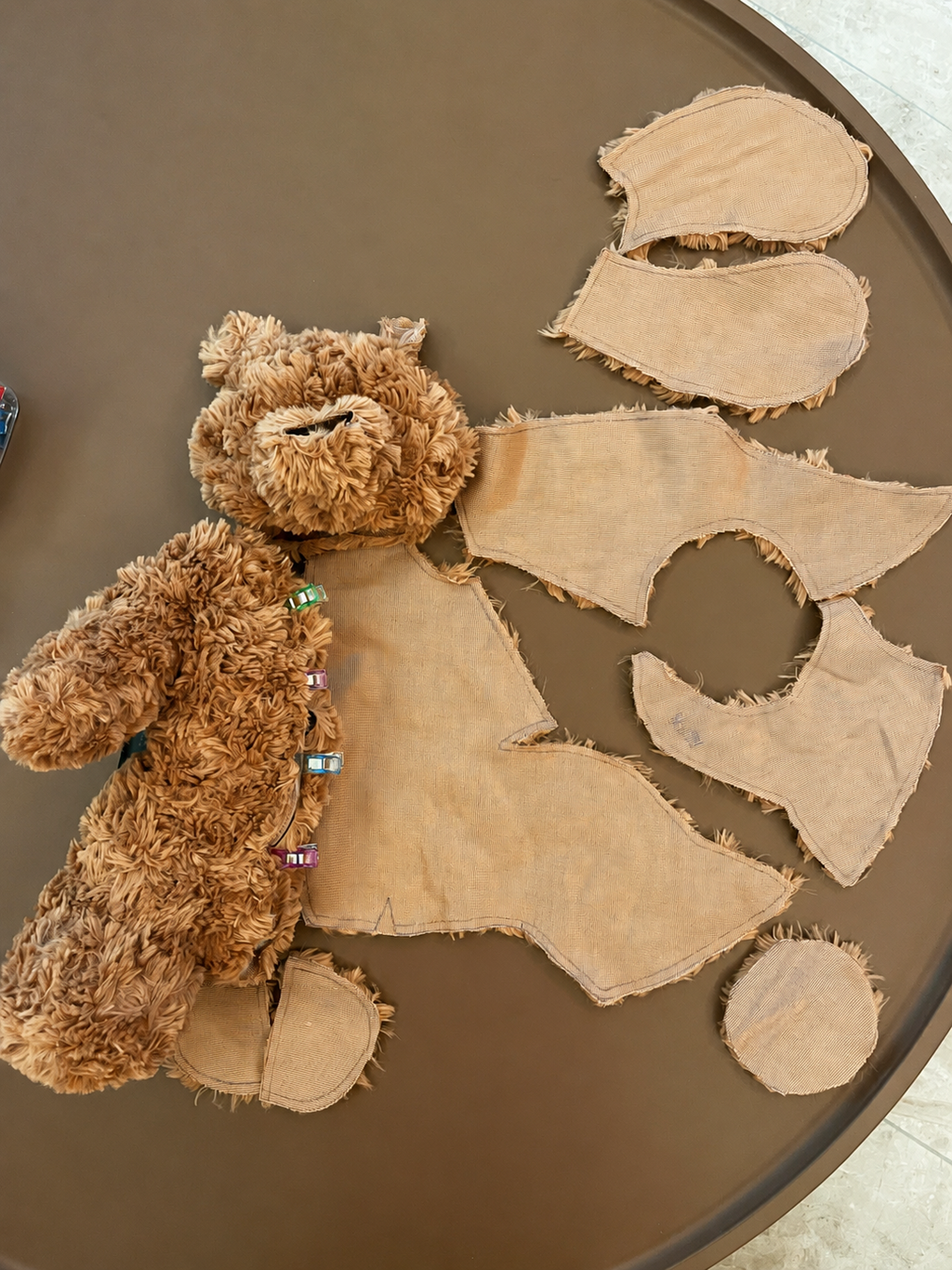 A teddy bear sewing pattern and making process, brown faux fur pattern and tan fabric pieces laid out on a round brown table. The bear has colorful beads around its neck, and several cut-out fabric body panels are arranged beside it.