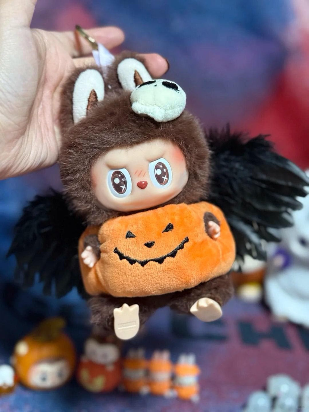 Halloween-themed labubu with a pumpkin and bat wings held by a hand.