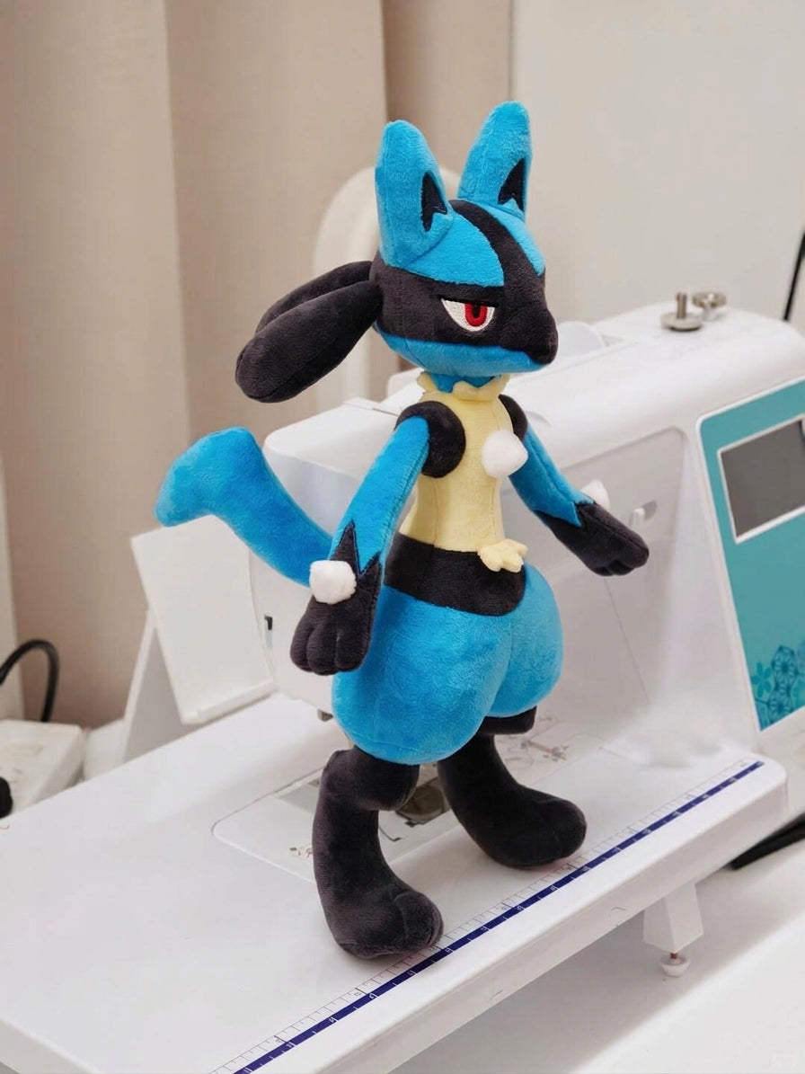 A blue and black Lucario plush toy character with red eyes stands on a white sewing machine in an indoor setting.
