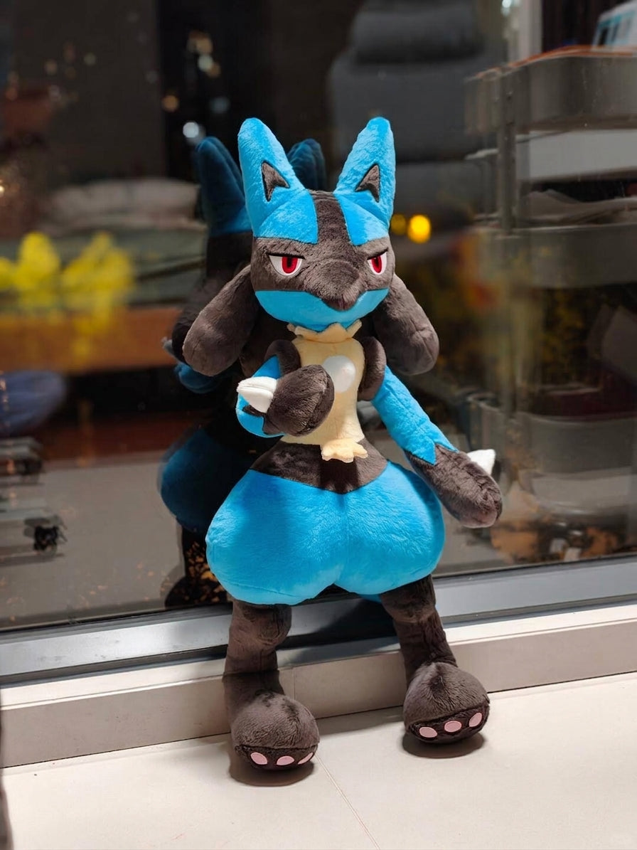 A blue and dark gray lucario plush toy resembling Lucario, posed sitting on a windowsill with a blurred indoor background visible behind it.