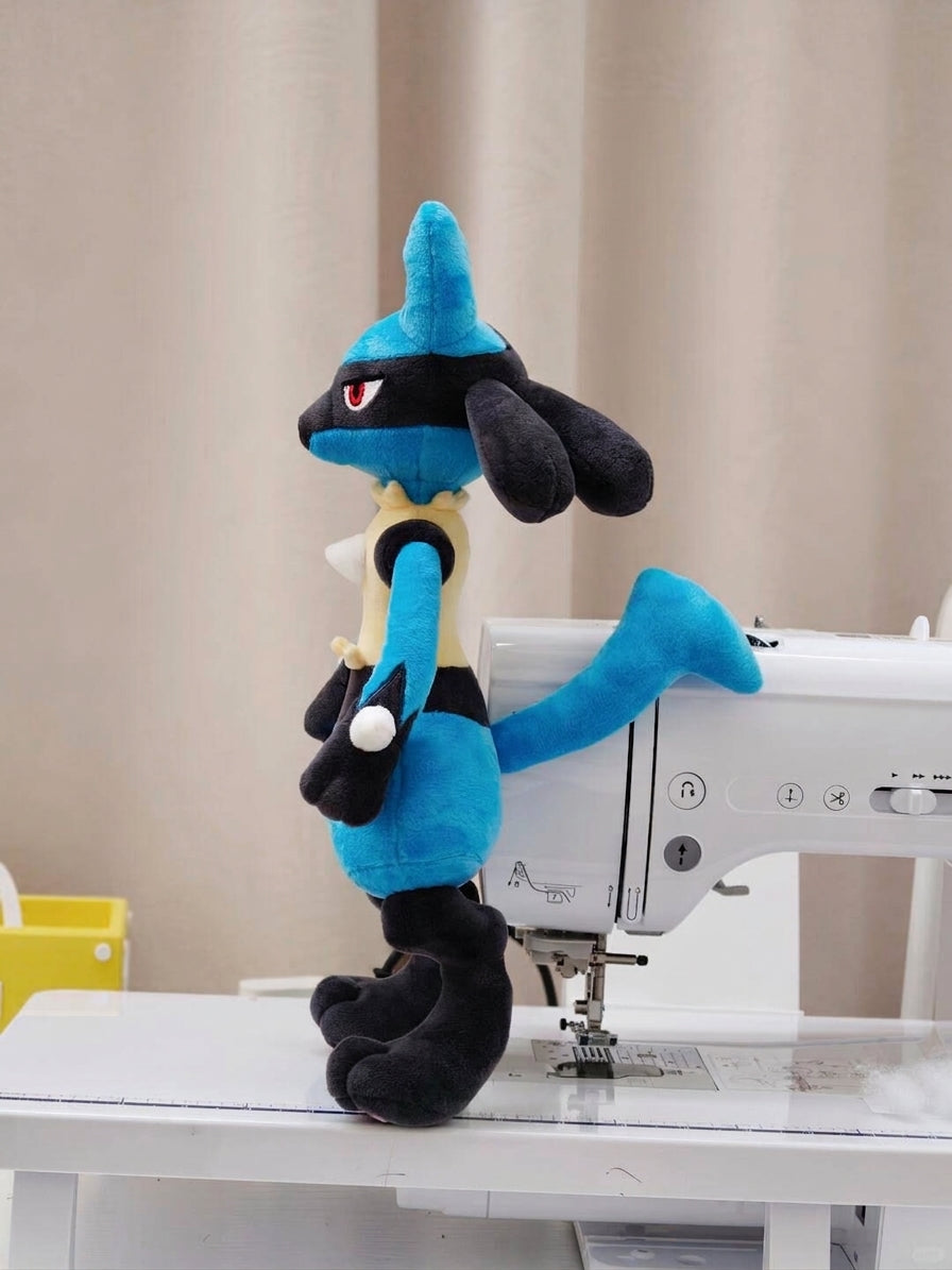 A blue and black lucario plush toy standing on a white sewing machine table in front of a beige curtain background.