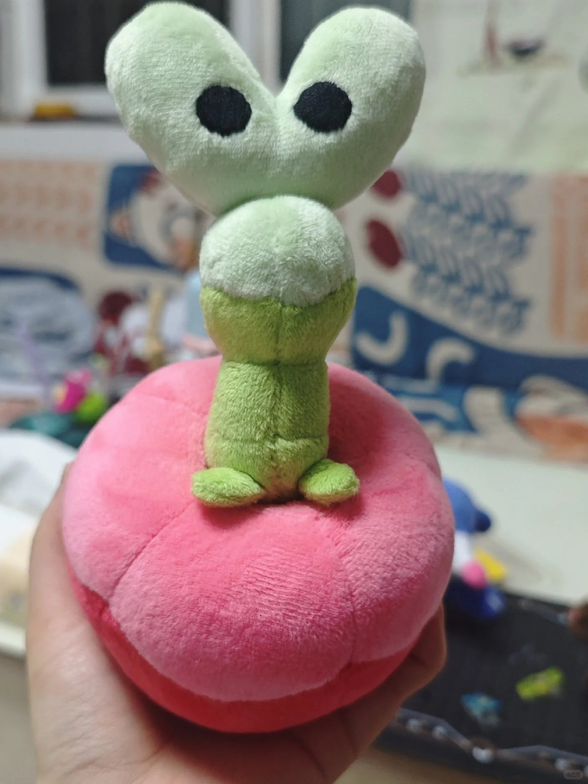 A Applin plush toy featuring a light green frog-like character with large black eyes sitting on a pink mushroom cap, held in a hand against a blurred indoor background.