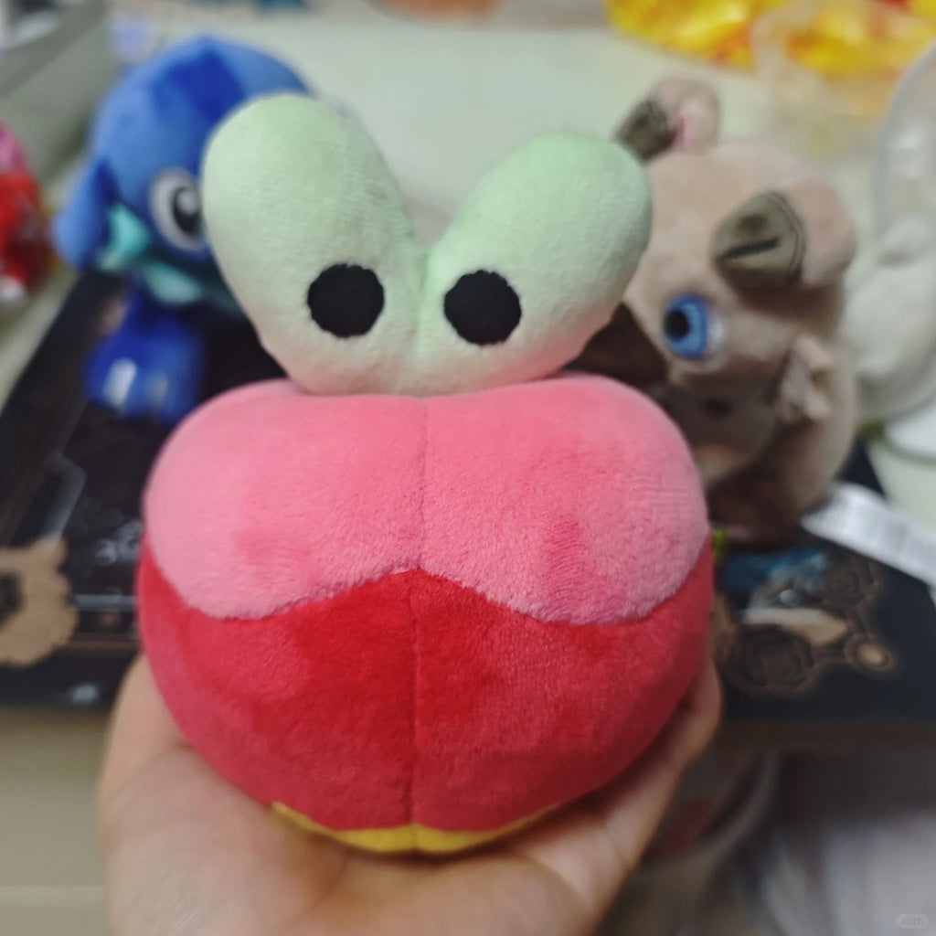A small Applin  plush toy held in a hand, featuring a pink and red body with two large light green eyes on top and a small yellow accent at the bottom.