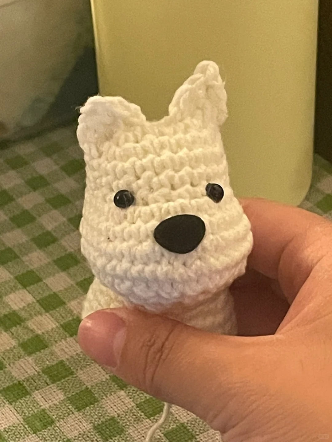 White crochet dog toy held in a hand with a green checkered tablecloth in the background