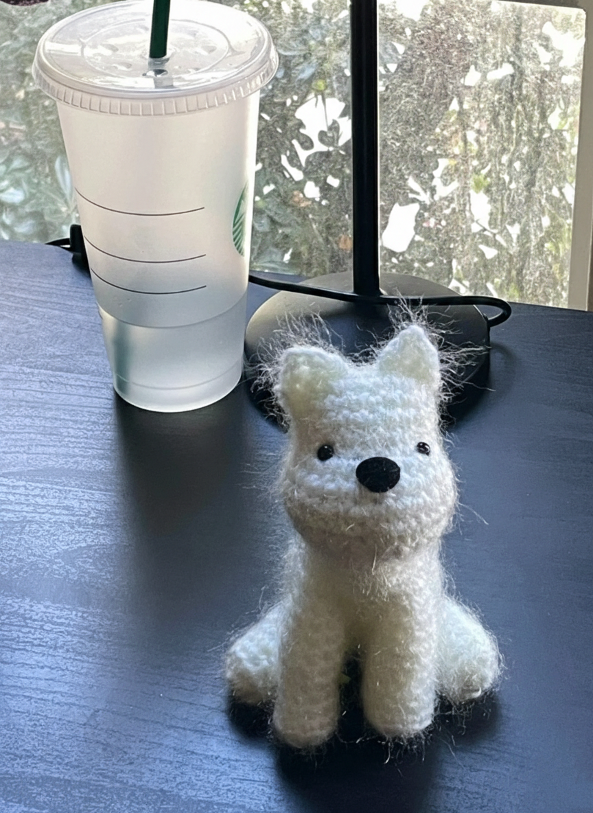 White knit crochet white dog toy next to a clear Starbucks cup on a dark surface with a window in the background.