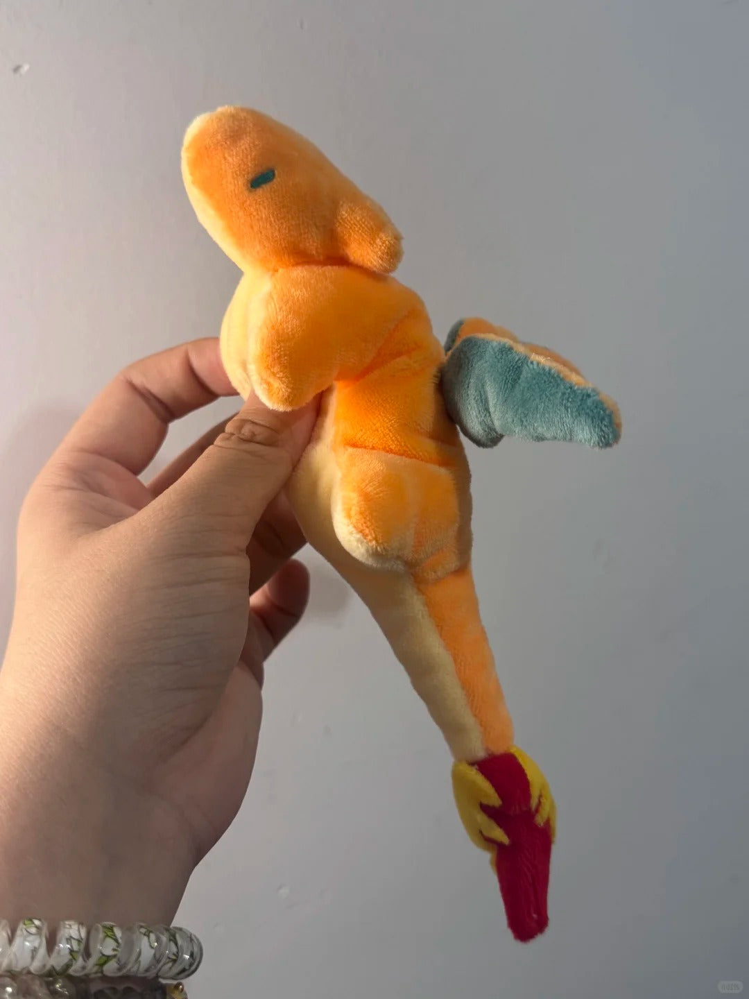 A small plush toy held in a person's hand against a plain light gray wall. The toy is orange with a blue wing, yellow accents, and a red tail flame.