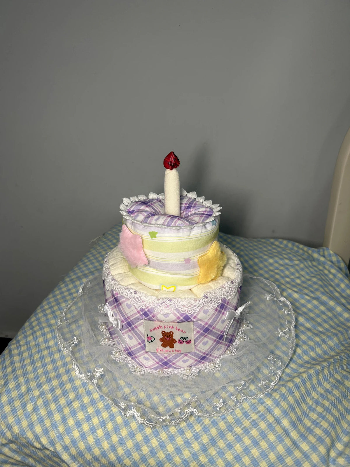 A diaper cake styled like a two-tiered birthday cake, made from folded diapers with purple plaid wrapping, white lace trim, pastel accents, and a small white candle topped with a red jewel, displayed on a clear plastic stand over a blue-and-white checkered blanket.