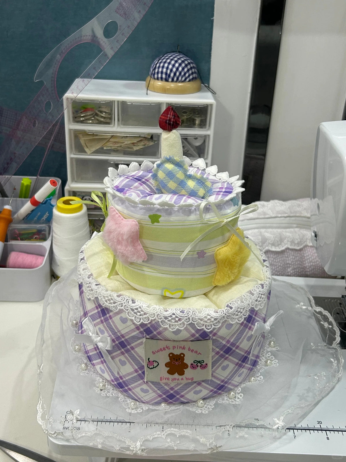 A handmade baby shower cake made from stacked diapers, decorated with a purple and white checkered base, white lace trim, pastel green and yellow stripes, and a small white candle on top, displayed on a sewing table with sewing supplies in the background.