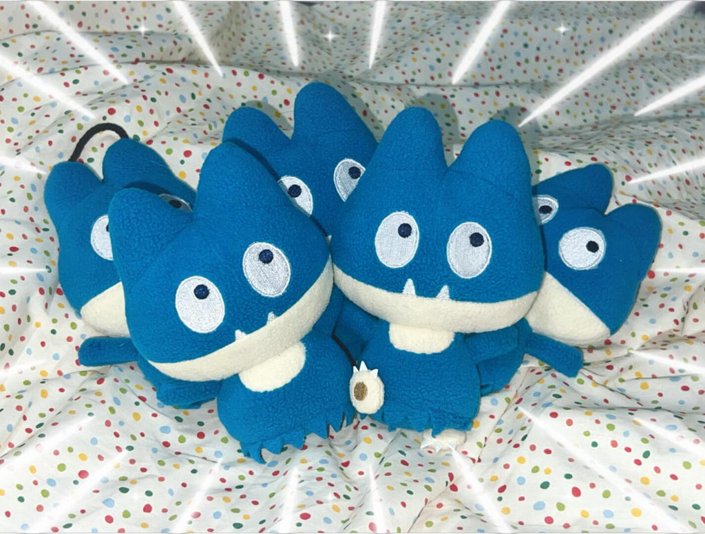 A group of blue plush snorlax monster keychains with large white eyes and small white teeth, arranged together on a white polka-dot fabric background with a digital starburst overlay.