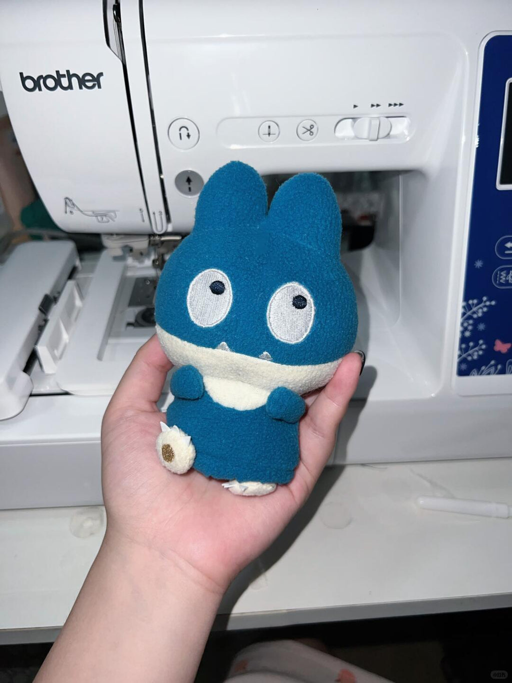 A blue and white Pokemon snorlax plush toy character held in a person's hand in front of a white Brother sewing machine.